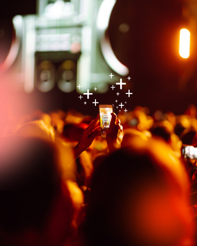 A crowd at a live show, one person holding up a phone as glowing plus marks drift above it