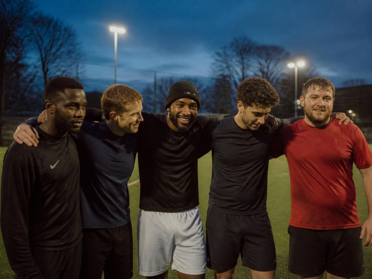 A five-a-side football team gathered on a floodlit pitch at dusk
